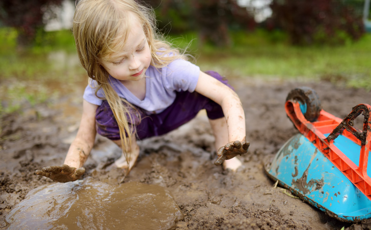 Funny Little Girl Playing In A Large Wet Mud Puddle On Sunny Summer Day. Child Getting Dirty While Digging In Muddy Soil.
